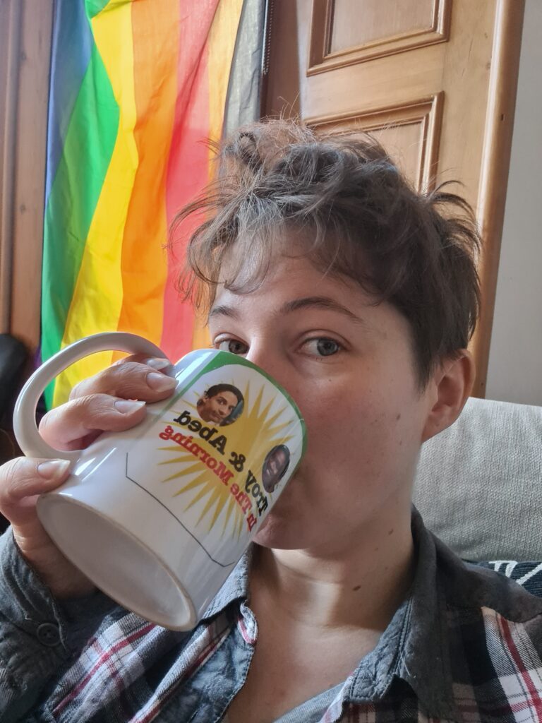 Photo of a person with short brown hair and white skin sitting in front of a pride flag and drinking coffee out of a 'Troy and Abed in the morning' mug