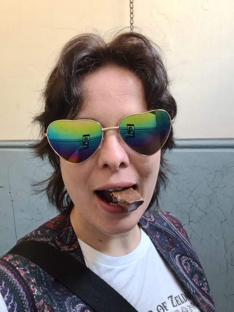 A person with brown hair and white skin wearing rainbow-coloured heart-shaped sunglasses and eating a chunk of milk chocolate